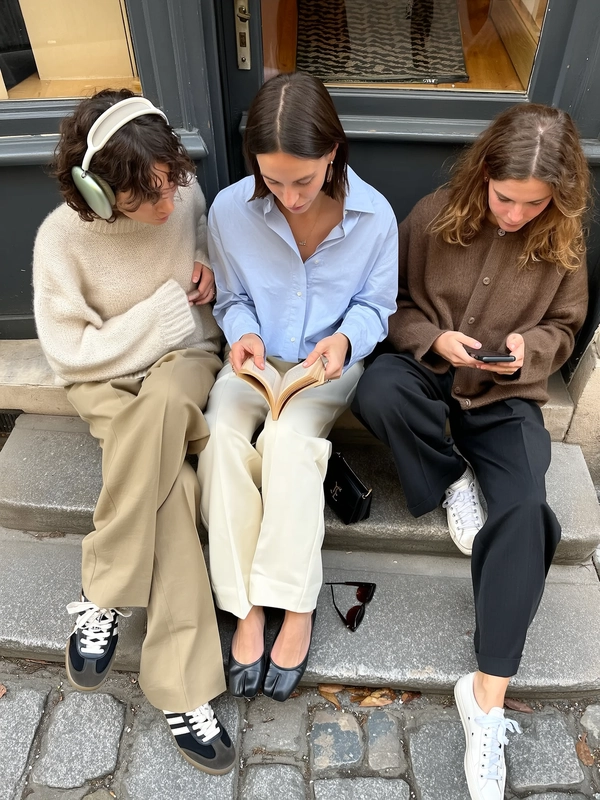 A candid iPhone-styled photo capturing a stylish trio casually seated together on worn stone steps outside a boutique on a quiet Parisian street. The first woman wears a soft cream UNIQLO x MARNI oversized knit sweater paired with tapered beige RECTO trousers and vintage-inspired Veja sneakers, large over-ear headphones resting lightly on her softly curled short hair. The second sports a loose-fitting pale blue Lemaire shirt layered over off-white tapered Our Legacy trousers and elegant The Row Ava Flats, intently reading a small paperback book, her straight shoulder-length hair tucked behind ears under minimal natural makeup, while a discreet Céline Phoebe Philo era Box Bag rests beside her. The third casually scrolls her phone wearing a relaxed-fit brown blurhms cardigan, loose black COS cropped pants, and classic white Converse, her tousled medium-length hair catching gentle daylight. Natural soft daylight softly illuminates their skin revealing pores and subtle imperfections, casting gentle shadows that enhance nuanced fabric textures, delicate creases, and visible fibers in their minimalist, high-quality clothing. The textured stone steps beneath them, an aged charcoal grey doorframe behind, and an uneven cobblestone street partially visible frame the scene authentically. Casual reflections shimmer subtly in sunglasses placed nearby. The composition features an intentionally informal slight tilt and off-center framing, captured from a casual overhead angle, conveying genuine, unposed intimacy and spontaneous Parisian street atmosphere true to candid iPhone photography aesthetics.