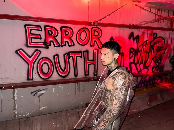 A candid urban iPhone photograph capturing a male model with abstract tattoos covering his arms and neck, piercing grey eyes fixed thoughtfully ahead. He stands inside an industrial loading bay bathed in pulsating red strobe lights, casting sharp shadows and warm reflections across the scene. Behind him, prominent layered stencil graffiti spells out large text reading:
“ERROR”
“YOUTH”
with overlapping cut-through letters creating raw street art depth. The model wears a transparent, slightly fluttering parka that catches the shifting light as if responding to the vivid graffiti. The concrete loading bay walls display weathered paint and subtle spray-paint drips. The framing is slightly off-center and tilted, with a spontaneous tight crop that imitates a casual, documentary-style street portrait shot on an iPhone. Textures of peeling paint, rough concrete, and layered graffiti tags add to gritty urban authenticity under moody red strobes reminiscent of candid nighttime street photography.