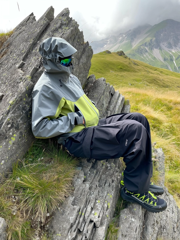 A casual, authentically candid photo spontaneously captured with an iPhone, featuring an individual reclining comfortably against rugged, textured rocky surfaces on a grassy alpine hillside. The person wears a two-tone slate gray and neon lime Arc'teryx hooded jacket with the hood pulled up, adjustable cords naturally loosened, the durable, waterproof fabric realistically creased with subtle dirt marks from mountain use. They sport loose-fitting black cargo pants, their relaxed posture showing natural fabric folds, complemented by practical neon lime-accented black sneakers. The individual's face is partially obscured by the hood and slightly turned away, wearing realistic reflective sunglasses catching diffused daylight. Softly diffused daylight filters through light clouds, casting gentle shadows over the textured rocky and grassy terrain, enhancing the natural mountain ambiance. The surrounding alpine meadow is dotted with wild grasses and jagged rock formations, distant mountain ridges faintly visible in misty light. The photo is framed with a casual, slightly tilted angle, emphasizing the relaxed, spontaneous intimacy and genuine outdoor authenticity typical of trendy gorpcore-style iPhone photography, with meticulous attention to the detailed textures of waterproof fabrics, natural terrain, and reflective lenses.