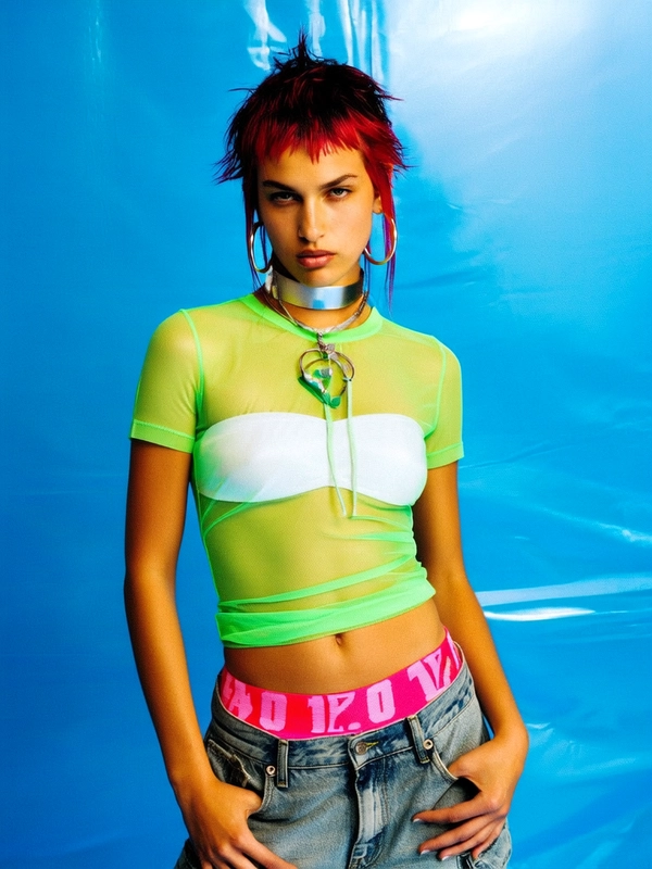 She's captured in a medium close-up against a glossy electric blue studio backdrop, the lighting casting soft shadows that accentuate the texture of her look. Wearing a sheer neon green mesh top layered over a plastic white bandeau, her styling insists on the raw Y2K aesthetic without explicit nudity. Her low-rise, bleached denim cargo pants slouch just enough to reveal a neon pink waistband with bold block lettering. Her hair is dyed a blazing cherry red, styled into sharp, spiked tufts with visible dark roots. Clashing with the transparent top, a thick silver choker and large hoop earrings add a subtle punk edge. Her gaze is direct, unflinching, carrying a cool detachment that fits the editorial mood perfectly—posed and still, demanding presence without softness. This captures the early-2000s digital camera vibe with its slightly skewed framing and grainy texture.
—casual candid early-2000s Y2K snapshot, grainy softness