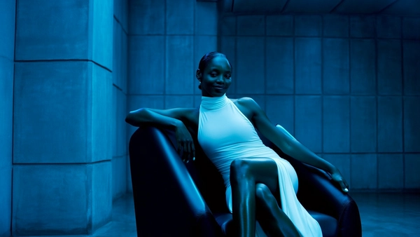 Cold, directional blue lighting cascades across a muted, industrial setting defined by gridded concrete walls that absorb shadows in deep, calming blues. At the center, a woman sits poised in an angular black chair, her legs crossed elegantly, radiating quiet command in a sleek, high-neck, sleeveless white dress that contrasts softly with her smooth skin and the stark environment. Her hair is pulled back tightly, revealing a poised, subtle smile that suggests quiet confidence and control. The framing is intimate and eye-level, emphasizing the taut tension between the woman’s relaxed yet magnetic presence and the austere, minimalist backdrop. The scene’s tactile richness emerges from the sheen on her legs against the matte roughness of the walls, the leather of the chair, and the delicate fabric of her dress flowing gently to the floor. This composition blends contemporary cinematic gloss with a cool, restrained color palette, evoking mystery and poised authority.