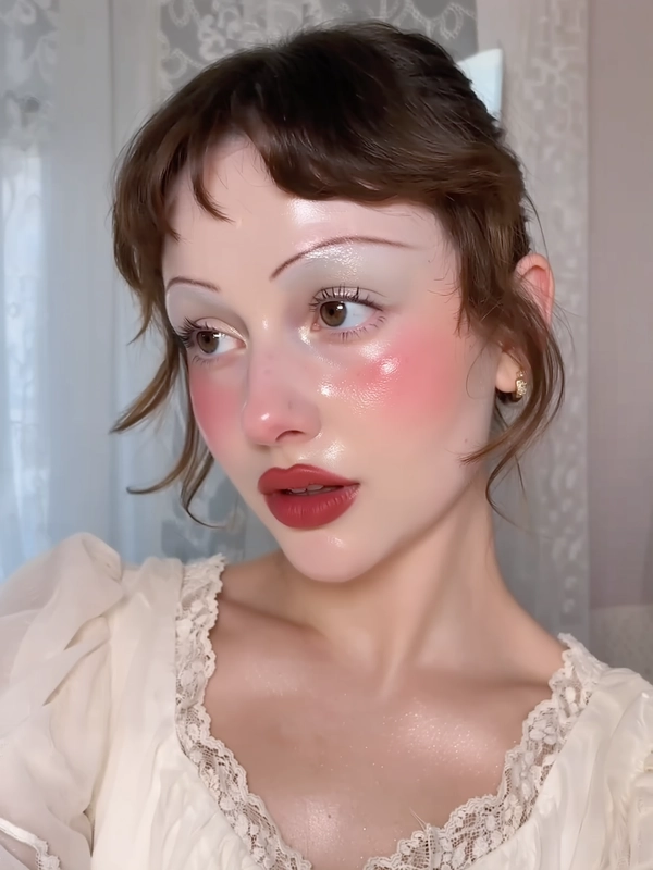 A close-up iPhone-style portrait of a young woman with retro finger waves softly framing her face, brunette hair with subtle shine and natural stray hairs visible. Her skin is creamy and luminous with a dewy finish that shows visible pores and natural texture. She wears stylized babydoll makeup featuring heavy pink blush extending across her cheeks and under her eyes, shimmering pastel eyeshadow in soft pearl white with subtle sparkle near the inner corners, and a matte red lip providing a bold but natural texture contrast. Her eyebrows are natural dark brown, brushed upward for a soft, natural look. The photo captures her from just above the head to below the collarbones, showing delicate sheer fabric in a pale cream blouse with puff sleeves and lace trim framing her shoulders and collarbone. The background softly blurs pale pastel tones with hints of textured indoor elements like curtains. Lighting is natural window daylight mixed with subtle iPhone flash, creating gentle highlights on her luminous skin and soft shadows under her chin. The composition is casual and slightly tilted, with the subject gazing just off-camera with a relaxed expression and parted lips. Fine details such as stray hairs, fabric weave, skin pores, and matte lip texture are clearly visible, enhancing the spontaneous intimacy and realistic texture of the image.