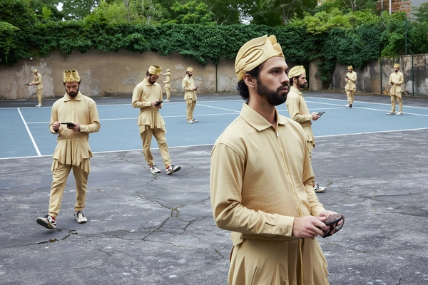 An imaginative, casually surreal snapshot featuring a man duplicated multiple times across an abandoned tennis court. He wears an all-beige layered outfit paired with a sculpted headpiece, identical in every copy. Each figure is calm yet tense, engaged in distinct tasks or gazing in various directions, forming a surreal choreography. The background shows overgrown fences and cracked asphalt, rendered with authentic textures of peeling paint, rusted metal, and weathered concrete. Natural daylight softly illuminates the scene, highlighting detailed fabric weaves, skin texture, and subtle imperfections. The composition employs creative angles and overlapping scales—from close-ups emphasizing facial calmness to distant figures sharply focusing on their tasks—capturing spontaneous candidness characteristic of iPhone photography and heightening the surreal repetition and interaction.