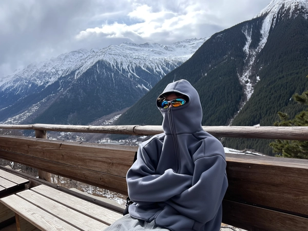 A spontaneous, candid iPhone photograph featuring a young person seated on a wooden terrace overlooking a vast alpine mountain range blanketed in snow. The individual is bundled in a slate gray oversized fleece hoodie with exaggerated seams and a large hood pulled firmly up, zipped up to cover the nose concealing most of their face, exuding a cozy cold-weather vibe. Their arms are tightly crossed in a humorous, dramatic pose, adding character to the silhouette against the soft snowy backdrop. Reflective sports sunglasses peek naturally from beneath the hood, their lenses catching diffused reflections of the moody overcast sky and snowy slopes. Wooden benches and a rustic railing frame the foreground, textured with weathered grain visible in the soft light. Surrounding dark green forests and patches of melting snow on alpine slopes provide a rich, textural contrast to the soft fleece fabric. The lighting is slightly diffused under a cloudy sky, emphasizing natural shadowing on fabric folds and wooden surfaces. The composition is casually framed with a gentle tilt, instilling natural intimacy and authenticity typical of trendy, candid winter mountain outdoor photography with a futuristic fashion edge.