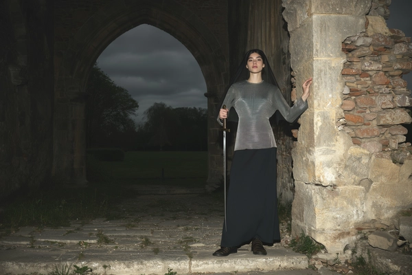 A wide, full-body, eye-level shot from an early 2000s-style digital camera explicitly capturing a young woman standing solemnly beneath a ruined stone archway in a dark, overcast evening setting. She wears a fine chainmail shirt layered over a long dark slipdress, her hair neatly pulled back with a thin veil resting gently on her shoulders. Her posture is upright and composed, one hand lightly tracing the cracks in the aged stone wall beside her, sword held vertically at her side. The harsh direct flash lighting characteristic of early digital cameras emphasizes the subtle sheen and imperfections of her metal armor, the delicate textures of her fabric dress, and natural skin details. The scene is filled with muted, desaturated tones and deep shadows, creating a moody, introspective atmosphere that blends medieval refinement with contemporary elegance. The framing is a thoughtful, elegantly balanced wide shot maintaining eye-level perspective to highlight her knightly dignity within this historic environment, perfectly embodying the medieval-core aesthetic with authentic early 2000s digital photographic qualities.