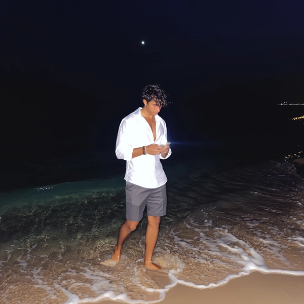 A sudden flicker of iPhone flash cuts through the night's deep blackness, revealing his relaxed stance where gentle waves whisper around bare feet sunk into cool sand. His crisp cotton-linen button-down shirt drifts ever so slightly with the seaside breeze, paired effortlessly with tailored shorts that suggest understated refinement. The textured shoreline glimmers faintly under the moon’s shy glow, while faint sheen on sun-kissed skin and the tousled crown of hair catch catch subtle light, embracing organic imperfection. Two-thirds of the frame stretches vast and velvety dark, offsetting his quiet poise, the softly blurred lapping surf echoing calm—flash-lit elegant beach snapshot, captured on iPhone.