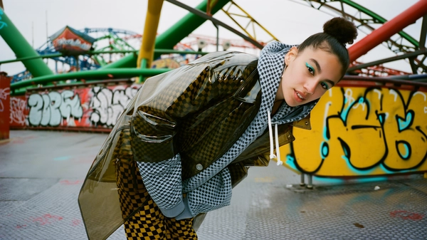 A spontaneous, candid image of a young woman standing near a graffiti-covered amusement park ride, wearing a glossy transparent coat layered over checkerboard print pants and an oversized mesh hoodie. Her hair is styled in a braided topknot, and she sports cyan eyeliner that subtly catches the natural daylight. The scene is lit by soft, diffused outdoor light that highlights the glossy sheen of the coat and the textured fabrics, alongside natural skin details and minimal makeup. The framing is informal and slightly tilted, capturing the vibrant, raw ambiance of the amusement park surroundings in a genuine, unposed moment characteristic of iPhone photography.