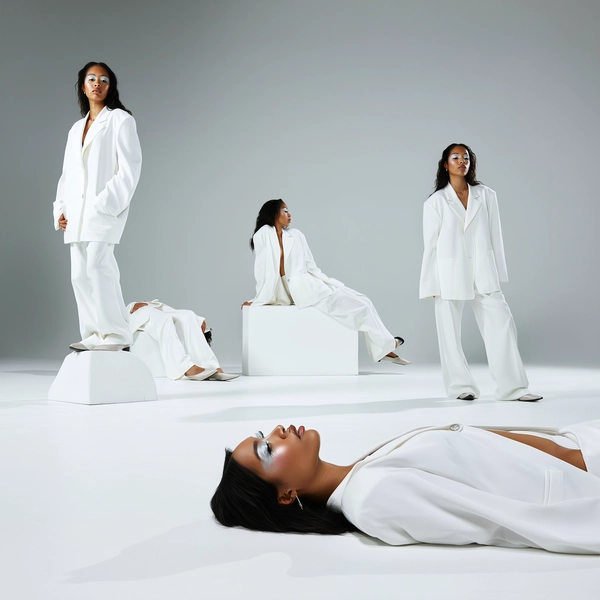 An imaginative, casually surreal snapshot featuring a woman duplicated numerous times, each instance clad in the same oversized white suit, her face adorned with glass-like makeup and bleached lashes. She appears in diverse calm and composed poses—standing, sitting, and laying—distributed naturally across minimalist white platforms within a starkly lit photo studio. Sharp, natural-like studio light highlights the intricate textures of her suit fabric, delicate makeup shimmer, bleached lashes, and subtle skin details. The composition employs creative angles and varied scales, with some figures closer and others receding, evoking a hyper-real yet spontaneous iPhone-captured moment brimming with surreal repetition and intimate authenticity.