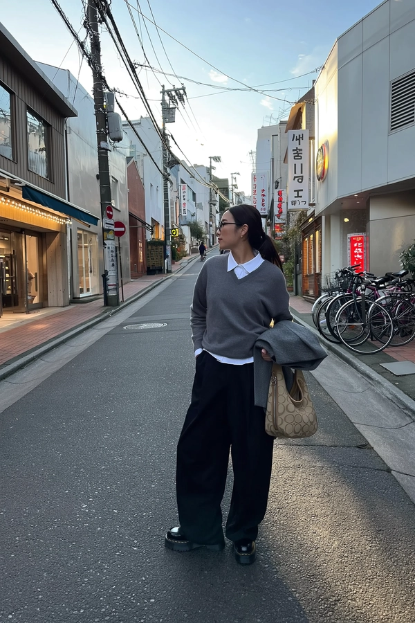A casually poised woman stands mid-step on a sun-dappled, gently sloping street near Koenji, her charcoal-gray V-neck sweater layered thoughtfully over a crisp white shirt, wide-leg black cargo pants skimming cracked pavement and her chunky black platform shoes catching faint neon reflections. Draped over one bent arm rests a folded gray coat, while a textured beige Coach Hobo bag hangs effortlessly by her side, its leather subtly creased under Tokyo’s muted afternoon light. Her hair is tied back neatly, round dark-rimmed glasses resting patiently as she gazes toward overhead electric cables tangled above narrow canal-side shops. Behind her, the quiet street climbs past faded kanji signages, occasional silhouettes of cyclists drifting by and a cluster of parked bicycles near a glossy konbini entrance. The iPhone’s handheld blur captures the pale blue twilight sky seeping gently through the crisscrossed wires, soft tungsten flares kissing the edges of her silhouette. The honest scene breathes a peaceful urban rhythm, textured with scuffed asphalt grit and softly glowing shop fronts, painted with the casual grace of Tokyo’s everyday streetwear cadence. —Tokyo street snap, shot on iPhone