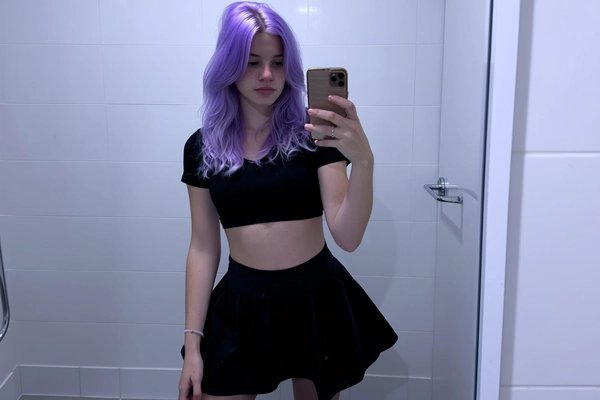 A candid mirror selfie of a young woman with vibrantly colored lavender hair styled in loose waves, dressed in a high-waisted black skater skirt and a fitted black crop top typical of mid-2010s fashion. She stands in a tiled bathroom with flat, clinical pure white lighting that creates minimal depth and no filters, evoking the raw, intentionally muted and color-drained Tumblr-style aesthetic. The image displays intentionally muted saturation and softly lowered contrast with subtly cool-toned shadows in pale lavender-gray hues. Visible textures include detailed skin imperfections, fine strands of dyed hair, and fabric grain of the skirt and crop top. Framing is casual, slightly off-center, and informally cropped, reminiscent of a spontaneous, unposed snapshot from mid-2010s youth culture photography.