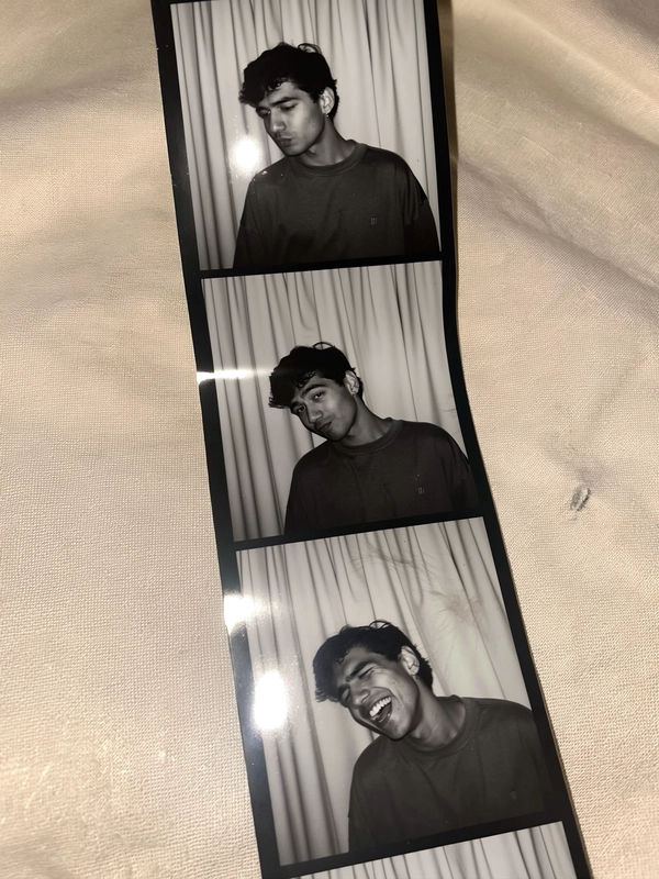 A vertical photostrip lies askew against a soft beige linen surface, slightly creased and bearing faint fingerprint marks. The black-and-white frames portray a young man in a loose long-sleeve tee against a textured neutral curtain background. In the first, he shrugs subtly, eyes caught in a quiet question; the next captures a playful head tilt, a hint of a smile flickering; the third erupts into a genuine, heartfelt laugh. Gentle iPhone flash reflections and muted contrast accompany the visible grain and soft natural shadows, with slight blur accentuating candid spontaneity. The strip’s casual tilt and the tactile details evoke the intimate nostalgia of authentic photobooth snapshots—contemporary photobooth snapshot, shot on iPhone.