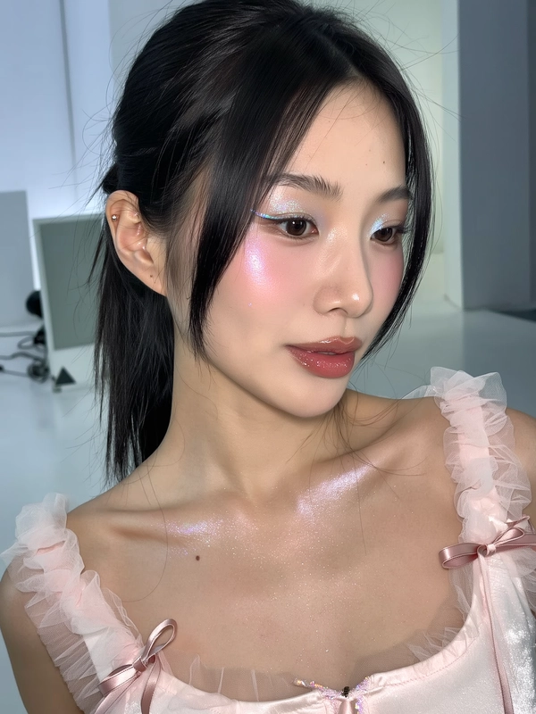 A close-up iPhone-style portrait of an East Asian woman with jet black hair tied in a sleek low ponytail, strands softly framing her face. Her translucent skin showcases a natural, dewy glow with visible pores and a subtle sheen along her nose, cheeks, and forehead. She wears holographic silver eyeliner that catches light with a delicate shimmer, paired with minimal pink blush softly diffused just on the apples of her cheeks, enhancing a fresh, youthful look. Her lips are coated in a high-gloss soft pink gloss with a tactile shine reflecting ambient light. Her eyebrows are a natural black, brushed upward gently without bleaching, complementing her expressive, relaxed gaze directed slightly off-camera. The composition is a tight upper-body framing from just above her head to just below her collarbones, captured at a casual, slightly tilted angle suggesting spontaneity. Her clothing features delicate babydoll-inspired details in soft pastel tones with sheer, textured fabric around the shoulders and a small ribbon bow at the neckline, subtly visible but not overpowering the composition. The background is a softly blurred futuristic studio space with muted, cool-toned hues of pale gray and silver, unobtrusive and supporting the subject’s presence. Lighting blends soft natural indoor ambient light with gentle phone flash, producing realistic shadows and subtle highlights across her face and collarbone. Fine textures like individual hair strands, gloss reflections, fabric weave, and skin details are clearly visible, all contributing to an intimate, authentic iPhone photo aesthetic.