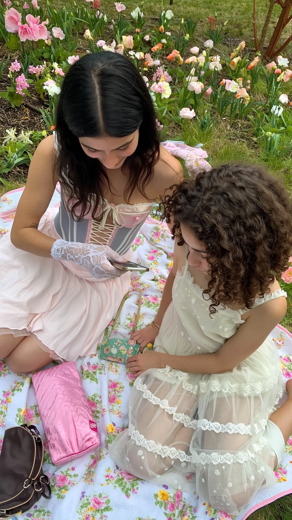 A softly intimate iPhone moment capturing two young women seated on a pastel-hued picnic blanket amid a blooming garden, dressed exclusively in whimsical Jenny Fax designs. One wears a delicate corset top adorned with intricate lace gloves, her long black hair falling gently around her shoulders in soft waves. The other dons a light babydoll dress featuring layers of sheer tulle and subtle ruffles, her curly brown hair framing a quietly reflective expression. Scattered pastel flowers enhance the garden’s timeless romance, while gentle daylight diffuses warmly, revealing the airy transparency of tulle, lace intricacies, and finely embroidered details. The scene is framed slightly tilted and elevated, balancing playful femininity with relaxed grace, the worn textures of the garden and soft color palette echoing Jenny Fax’s distinctive vintage-inspired delicacy.