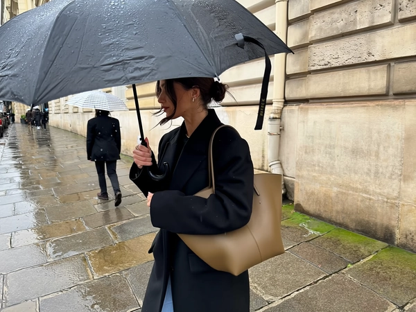She strides close to a beige stone façade, her sleek black coat crisp and structured, olive-brown leather shoulder bag tucked warmly beneath her arm, dark hair pulled into a loose low bun with moisture-softened tendrils tracing her cheek. A large black umbrella speckled with water beads arcs above her, each droplet sharply etched in the soft overcast light that diffuses through cloud-heavy sky. Her hand clasps the umbrella pole near her face as rain slicks the uneven pavement, the wet stones reflecting muted greys and faint hints of green moss. Behind her, a distant walker fades into blurred focus, translucent umbrella haloed by the damp air. The frame crops vertically, tight on her torso and thoughtful side profile, the stone wall filling the right border in a quiet, respectful embrace. Slight hand shake and lens shimmer lend candid intimacy, capturing the hushed mood and tactile rain textures with effortless elegance—rainy city snap, shot on iPhone