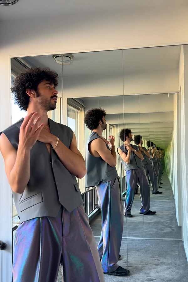An imaginative, casually surreal snapshot taken with an iPhone inside a mirrored hallway where a man is duplicated multiple times, always wearing the same sleeveless jacket and loose iridescent trousers. The scene features endless reflections multiplying the figures, each frozen mid-gesture—one with hand raised, another with back turned, and a third with an arm across his chest. Natural daylight softly illuminates the iridescent fabric, highlighting detailed skin textures, subtle hair strands, and authentic shadow play. The composition exploits surreal scale and perspective through overlapping reflections, framed intuitively with unexpected cropping to evoke an organic, spontaneous feel characteristic of casual iPhone photography.