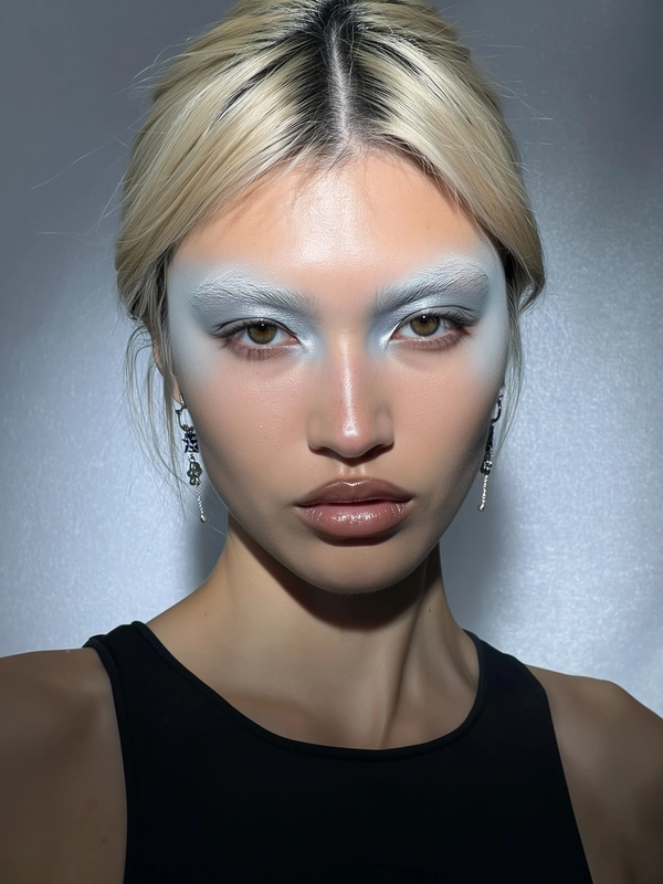 A close-up iPhone selfie portrait of a young woman with a sharp jawline and naturally bleached eyebrows that softly blend into her skin, creating a pale blonde subtle effect. She wears airbrushed metallic makeup with muted silver and icy tones around her eyes and softly glossy nude lips, capturing a sophisticated yet natural metallic sheen. Her expression is neutral and calm, directly engaging the camera with realistic skin textures highlighted despite the cold, harsh studio lighting that creates crisp shadows and reflections. She is styled simply with a minimal black top and delicate silver earrings. The background is matte gray with a softly textured, neutral finish, emphasizing the cold studio atmosphere while preserving the effortless, candid vibe typical of authentic iPhone selfies.