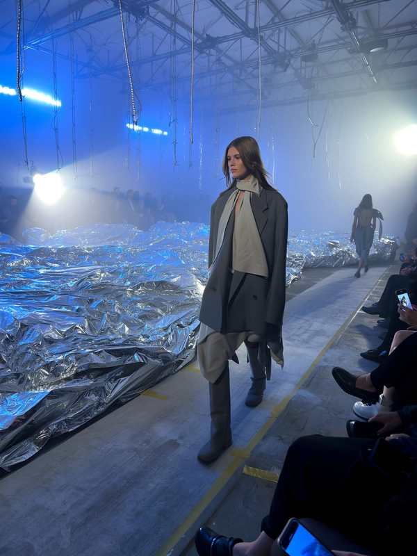 A model draped in layered muted greys and soft taupes crosses a surreal set strewn with crumpled silver metallic sheets catching reflections like moon craters. Her silhouette is relaxed yet precise, with oversized asymmetric tailoring brushing against thigh-high matte boots. Soft, cool blue backlights wash the industrial space into dreamlike hues, while sharp white spotlights carve subtle shadows over her calm, focused expression. The runway, a narrow strip of worn concrete, shows faint taped lines and slightly scuffed marks from heavy boots, blending tactile grit into the ethereal scene. Guests sit quietly along low benches, their blurred outlines softened by the dreamy haze as a few phones raise in the dim glow, blue light pooling against bare steel trusses overhead. The iPhone lens picks up gentle motion blur on the swinging metallic cords dangling from the ceiling, sensor noise in the deep shadows, and a delicate bloom around the soft LEDs that fill the space. Snap taken from an oblique front-row angle, with a slight hand-held shake and an intimate tight crop that draws you inside the lunar dreamscape—contemporary runway snapshot, shot on iPhone