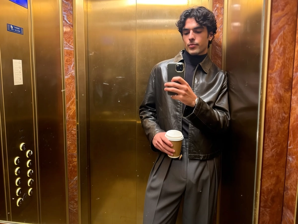 Bathed in the golden haze of vintage brass and faded marble panels, he leans back slightly in the warmth of the old-school lift, a dark turtleneck snug beneath a cropped leather jacket that catches the soft glow like worn velvet. His pleated trousers fall in fluid, relaxed folds, the fine weave faintly outlined beneath the flickering light, while a coffee cup balances effortlessly in one hand, the other holding the phone chest-high in a classic elevator mirror moment. Fingerprint smudges mar the glass just enough to soften the reflection, and his gaze stays fixed on the screen rather than the lens, an unbothered, almost knowing air about him as the aged brass buttons blink with quiet life beside him. The subtle grain of the iPhone snap settles across the scene, the gently worn textures and warm hues embedding a calm, refined confidence in the frame. —elevator mirror capture, urban chic on iPhone