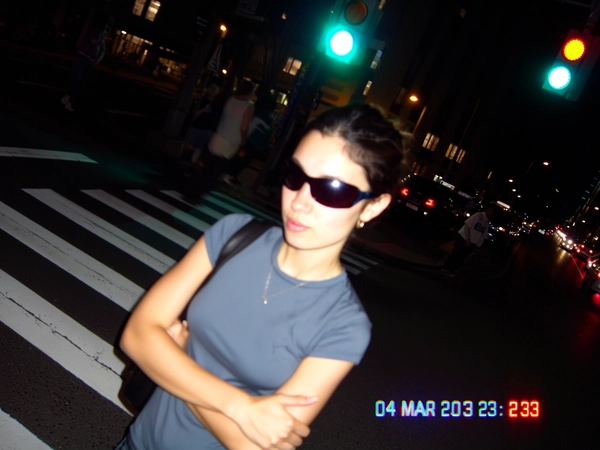The harsh tungsten glow spills unevenly over a woman standing at the crosswalk, her tight tee slightly faded by the digital sensor's soft focus edges. Her oversized sunglasses catch pixel fringing from the streetlight, which hovers as a glowing blur above her crossed arms. The moment blinks with on-camera flash bloom despite the night, while CCD noise speckles the shadows creeping around her silhouette. The view tilts diagonally, as if quick-zoomed without care, cropping the bustling background and featuring a jittery timestamp stamp: “04 MAR 2003  23:18” in ghostly blue. JPEG blocks crunch subtly near the traffic light, and a flickering battery icon stutters in the corner. The photo quality suggests a slightly grainy or low-resolution digital look, capturing a candid moment.—early-2000s digicam snapshot, grainy low-res quality