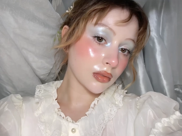 A close-up iPhone-style portrait of a young woman with soft, natural hair loosely framing her face, featuring subtle flyaways for texture. Her skin glows with a flawless, dewy glass-like finish that reflects a delicate pearl-white shine across her cheeks, nose, and forehead. Her makeup embodies an ethereal babydoll style with shimmering pastel metallic eyeshadows in soft silver and pearl tones, applied gently over the lids with a subtle blend into icy white near the inner corners. Heavy pink blush softly diffuses beneath her eyes and across the tops of her cheeks, adding warmth to the metallic hues. Her lips are coated with a glossy pale peach pink gloss that gleams gently under natural daylight. Her brows are natural-colored, light brown, brushed upward lightly without bleaching. She wears a vintage-inspired puff-sleeve blouse in a soft pastel cream with slight sheen and delicate lace collar featuring tiny pearl buttons and a subtle ruffle trim at the neckline. The sheer fabric layers catch the light, emphasizing texture and translucence near her collarbone and shoulders, which are softly lit with natural window light mixed with minimal phone flash. The composition is tightly framed from just above her head to below her collarbones at a casual, slightly tilted angle, with the woman looking softly off-camera in a relaxed, unposed expression. The background is softly blurred in pastel tones of pale silver and cream, with faint textures of fabric suggesting indoor setting. Fine details like visible skin pores, gloss texture, shimmer particles on the eyelids, delicate flyaway hairs, and realistic fabric stitching are clearly visible, creating a genuine, intimate, and spontaneous iPhone photo aesthetic.