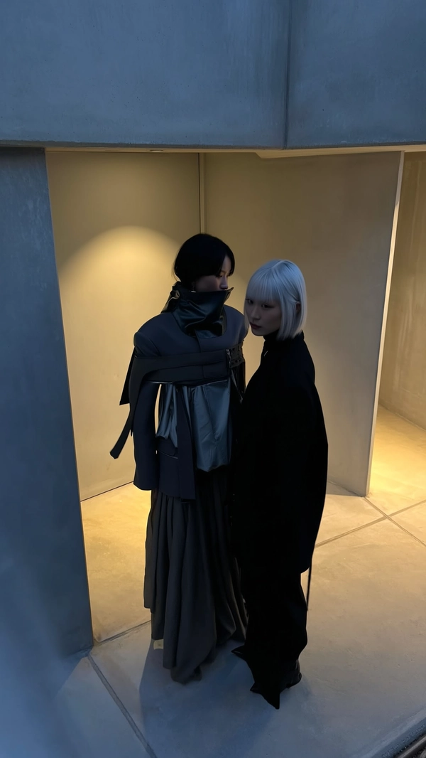 A softly dim-lit, dusk-inspired iPhone snapshot of two female models standing back-to-back near a gently illuminated muted grey Brutalist concrete structure. The taller model sports a layered, avant-garde Julius ensemble comprising a high-neck jacket partially obscuring the face paired with a long, textured lower garment, while the shorter figure's sharp features and silver hair subtly contrast the refined dark fabrics. Soft, diffused twilight light casts understated shadows across the matte textiles and angular concrete forms, with generous negative space and a slightly elevated, tilted downward viewpoint. The scene is framed by soft foreground concrete textures, evoking quiet intimacy and relaxed elegance without direct sunlight, harmoniously blending the avant-garde fashion with quietly moody Brutalist environs.