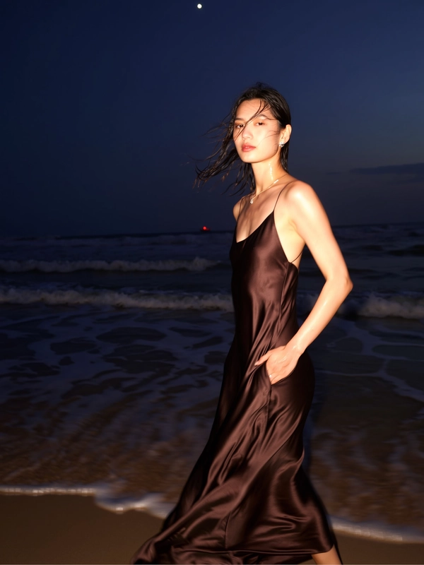 A sudden flash reveals her serene figure framed by softly undulating waves and dark sand, one hand tucked casually into the pocket of a flowing deep brown satin dress that ripples gently in the seaside breeze. The faint shimmer of her skin and damp tendrils of hair catch the subtle moon-glimmers while expansive night envelops two-thirds of the frame in velvety darkness. Her poised stance stands crisply against the blurred swirl of the hem, a quiet magnetism rising from the interplay of fluid fabric and natural textures, inviting calm admiration beneath the soft glow of distant ambient light. —flash-lit elegant beach snapshot, captured on iPhone.