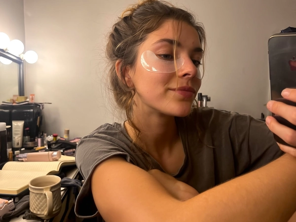 A young woman sits comfortably in a cozy makeup room, visibly wearing translucent hydrogel under-eye patches that gently hug the skin beneath her eyes. Her hair is loosely styled, some strands casually falling across her face, while soft, warm makeup room lights softly illuminate her smooth complexion and the texture of the patches. She wears a relaxed, casual top and slightly rumpled surroundings hint at a lived-in space, with an open notebook and a ceramic mug of tea resting nearby on a cluttered vanity. The selfie is taken with a natural front-facing iPhone angle capturing her face and upper body in a candid and intimate moment of self-care—skincare selfie, shot on iPhone.
