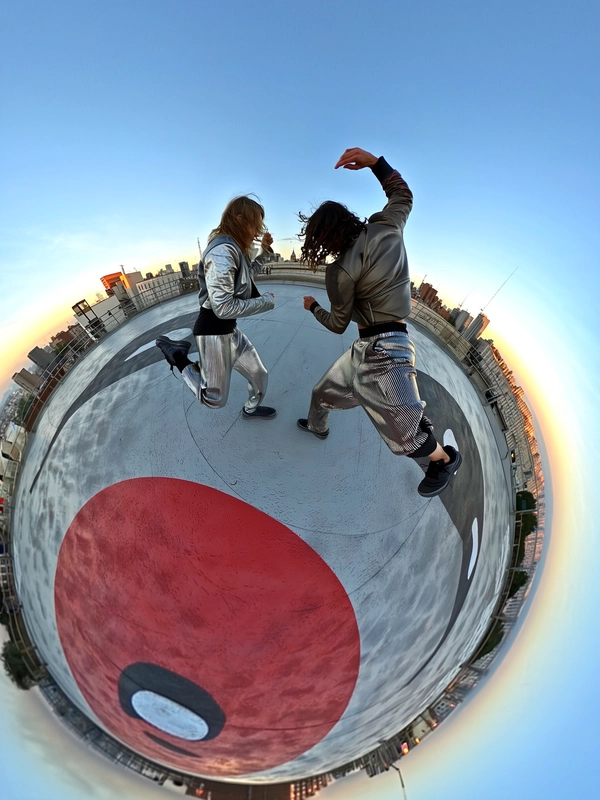 A spontaneous stylish 360-degree candid photograph set on a wide urban rooftop helipad featuring a large pop-art face painted boldly across the surface. Two dancers, captured mid-motion with natural, fluid poses, wear striking metallic pleated pants that shimmer with finely detailed reflective textures and cropped futurist jackets combining sleek, structured fabrics in muted silver and gunmetal tones. Their footwear is minimalist and edgy, with matte black sneakers showing subtle everyday wear. Their hairstyles are effortlessly tousled, conveying dynamic movement and natural texture. The setting includes the painted helipad's tactile concrete surface and the surrounding skyline faintly visible under soft natural daylight filtering through a clear blue sky, casting diffuse, authentic shadows enhancing the metallic fabric highlights and the bold graphic beneath. The image is taken with a spherical 360-degree camera positioned at ground-level center, angled upward, creating an immersive circular tunnel-like perspective that artistically distorts the rooftop's expansive space and the dancers' poses. This composition perfectly evokes the genuine, stylish spontaneity, tactile realism, subtle imperfections, and intimate urban energy distinctive of upward-facing 360-degree candid photography captured with a portable 360-degree camera paired with an iPhone.