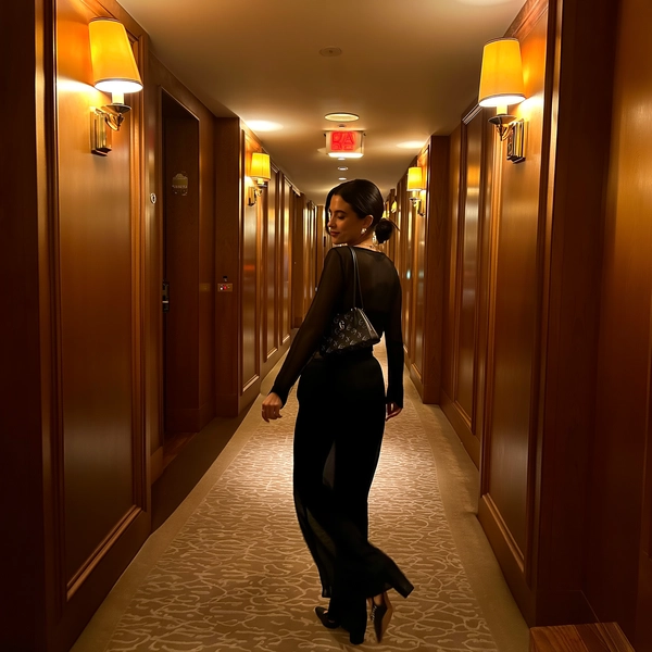 In a dimly lit hotel corridor clad with warm wood panels and bathed in symmetrical amber sconces, she strides with quiet command, caught mid-turn glancing back over her shoulder. Her sheer black top gently hugs her silhouette, delicately revealing skin beneath translucent fabric, paired with flowing high-waisted trousers that sway effortlessly with each step. The patterned plush carpet beneath her heels draws the eye forward like a runway, while her sleek monogrammed handbag hangs gracefully from one hand, the other swinging loose and natural at her side. The ambient glow softens highlights on polished brass fixtures, casting cozy shadows across textured walls. The moment hums with late-night hush, a private luxury stillness thick in the air, captured with subtle sensor grain and a soft-focus edge on the iPhone, lending this cinematic passage an intimate, unhurried glamour. —leisure-suite snapshot, shot on iPhone