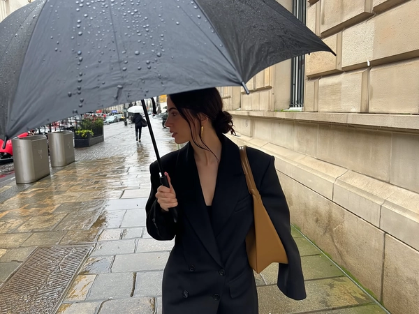 She strides close to a beige stone façade, her sleek black coat crisp and structured, olive-brown leather shoulder bag tucked warmly beneath her arm, dark hair pulled into a loose low bun with moisture-softened tendrils tracing her cheek. A large black umbrella speckled with water beads arcs above her, each droplet sharply etched in the soft overcast light that diffuses through cloud-heavy sky. Her hand clasps the umbrella pole near her face as rain slicks the uneven pavement, the wet stones reflecting muted greys and faint hints of green moss. Behind her, a distant walker fades into blurred focus, translucent umbrella haloed by the damp air. The frame crops vertically, tight on her torso and thoughtful side profile, the stone wall filling the right border in a quiet, respectful embrace. Slight hand shake and lens shimmer lend candid intimacy, capturing the hushed mood and tactile rain textures with effortless elegance—rainy city snap, shot on iPhone