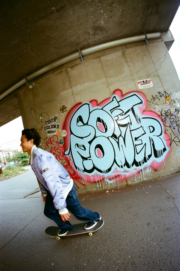 A spontaneous candid iPhone photo capturing a young man skateboarding past a graffiti-covered concrete underpass. He wears a pastel-colored bomber jacket paired with casual streetwear, his mid-motion blur echoing trailing drips of fresh spray paint. The graffiti prominently features large stylized text reading:
“SOFT
POWER”
with paint drips seeping into the cracks of the rough concrete wall. Surrounding the primary text are smaller, faded tags and layered stickers including iconic smiley faces and cartoonish figures slightly peeling at the edges. Natural diffuse daylight filters through the underpass, casting realistic soft shadows and enhancing the gritty textured surfaces of weathered concrete and paint-splattered ground. The framing is unintentionally tilted and off-center, shot from a low eye-level angle, embodying the raw street authenticity and spontaneous energy typical of iPhone documentary street photography.