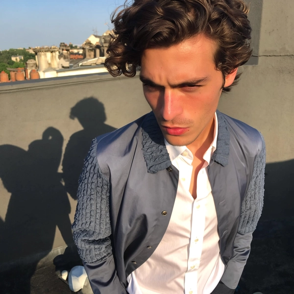 A spontaneous candid photo of a male model with sun-kissed skin and a focused, intense gaze standing casually on a rooftop with bold geometric shadows falling around him. He wears a slate-gray jacket featuring distinct textured sleeves paired with a crisp white shirt buttoned up to the collar, showcasing realistic fabric grains and subtle wrinkles. The late afternoon natural light creates soft highlights and shadows on his skin and clothing, enhancing authentic texture and natural skin details. The framing is slightly tilted and off-center, capturing a genuine, relaxed posture typical of iPhone photography. The overall mood conveys raw, effortless style with natural urban architectural elements in the background, emphasizing an authentic and intimate atmosphere.