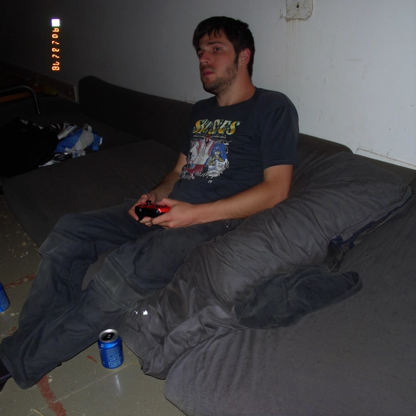 A sudden pop from the built-in flash washes over a man slouched on a threadbare couch, his graphic T-shirt catching a faint blur as he grips a game controller. Nearby, a dented soda can shimmers on the scuffed floor, its edges softened by low CCD sensitivity. The flash etches a harsh shadow onto the cracked paint of the wall behind, while faint pixel blocks jitter across the darkened folds of the fabric. The man's outline bleeds into the muted tungsten glow mixing with a hint of fluorescent overhead light, edges fuzzing with each tiny jitter of the handheld zoom. A slight tilt twists the scene off-kilter, the timestamp “04 MAR 2003  23:18” flickering in the corner alongside a half-drained battery icon. The photo quality is low-resolution and somewhat grainy.—early-2000s digicam snapshot, grainy low-res quality
