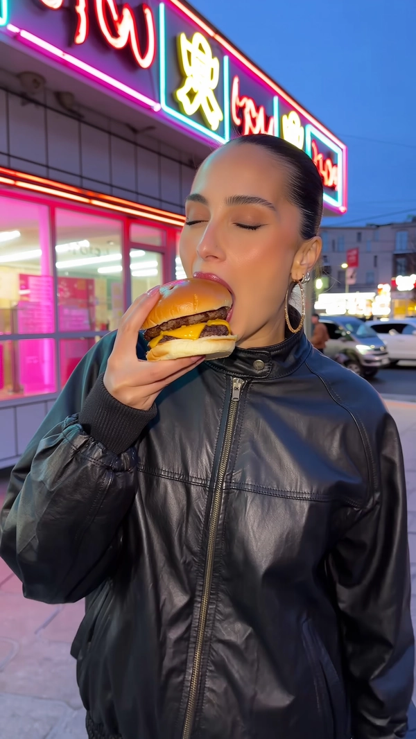 In the cool evening glow outside a neon-lit fast food joint, a striking woman in a smooth black leather jacket takes a deliberate bite of a stacked burger. Her hair, pulled back neatly with subtle makeup enhancing her flawless skin, frames her neutral, poised face. Her eyes close gently as the food touches her lips, a quiet ritual of satisfaction undisturbed by the vibrant signage flickering behind.

The burger’s glossy bun glistens under the neon’s electric pink and blue hues, while melting cheese stretches lazily from the patty, teasing the bite. A delicate sheen of lip gloss catches the light briefly as her mouth meets the food, the contrast of textures vivid against her calm expression. The cool air carries faint scents as her jacket’s leather catches scattered city light, each crease defined in sharp detail.

A casual iPhone angle captures the moment from just below eye level, highlighting the intensity of the eating gesture amid the busy urban backdrop. The subtle blur of passing cars and blurred signage meld into soft color washes in the distance, focusing attention tightly on her and the simple act.

This quiet, composed image melds streetwear edge with an editorial stillness, embodying the intimate precision of a fleeting, stylish bite — perfectly resembling a candid iPhone photo.