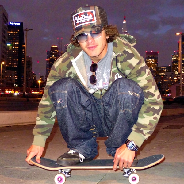 Crouched low on his skateboard under the neon-lit cityscape, he leans in close to the camera, the glow of blurred city lights framing his face in warm pastel hues. Sporting a baggy low-rise denim with patchwork details and a camo-colored zip-up hoodie layered over a slightly glittered graphic tee, his fitted Von Dutch cap sits tilted just right. Tinted sunglasses hang off the collar, and chunky DC skate shoes touch the pavement with casual ease. His wrist sports a chunky G-Shock watch, perfectly holding down that authentic Y2K vibe. The photo quality is low-resolution and somewhat grainy, capturing the raw energy of this spontaneous moment. The pixel-smearing and slight blur around the edges echo the soft glow of a late-night CCD camera shot. This close-up snapshot really nails that laid-back skater style from the early 2000s.—casual candid early-2000s Y2K snapshot, grainy low-res softness