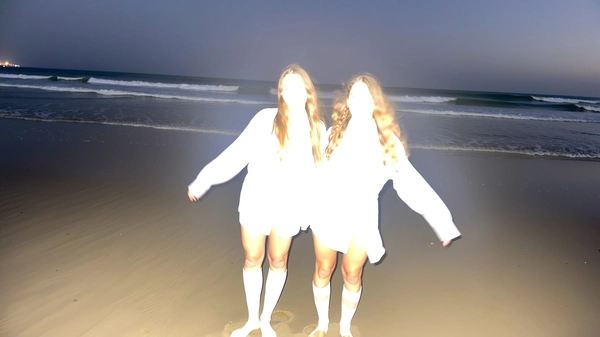 A spontaneous nighttime snapshot on a misty beach, captured with an intense flash from an iPhone. Two barefoot women with long hair stand side by side wearing oversized white shirts and long socks, their arms loosely linked. The intense flash overexposure completely bleaches out their faces and clothing details, leaving only ghostly, luminous outlines against the softly moonlit waves in the background. The imperfect framing and casual stance emphasize a surreal, candid moment.