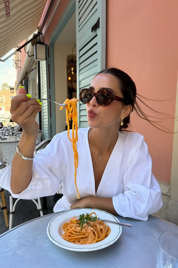 Basked in the soft late-morning glow of Capri's gentle sun, a striking woman settles at a small table outside a pastel-blue bistro, its colors melting into the warm Mediterranean air. Her hair is sleekly pinned back, and her oversized, retro tortoiseshell sunglasses shield soft, thoughtful eyes. She wears a lightweight white blouse with subtle embroidery, sleeves relaxed over slender arms, nails painted a vibrant neon yellow that catch fleeting reflections of the sky.

She lifts a fork elegantly twirled with glossy strands of fettuccine, coated in a silky lemon-cream sauce that gleams under the sunlight. The precise, fluid motion pauses just before her mouth, lips parted into a measured, serene gesture that holds a quiet poise. A delicate droplet of sauce clings briefly to her chin, adding an authentic undertone to the composed scene.

Around her, pastel hues and the warm limestone pavement create a serene, inviting backdrop, gently blurred by the arm-length vantage point of an iPhone snapshot. The breeze ruffles a stray strand of hair, while the delicate weave of her blouse fabric and the slight sheen of her lip gloss catch the light, weaving subtle texture into the frame.

Captured from a slightly tilted, casual angle as if by a nearby companion, the scene balances intimate detail with effortless style, embodying an editorial calm in Capri's sunlit charm, perfectly resembling a candid iPhone photo.