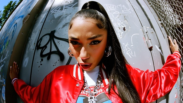 A wide shot of a woman with medium brown skin, detailed natural skin texture visible across her nose, cheeks, and forehead, and long sleek hair pulled into a high ponytail, captured in a middle close-up from a top-down wide-angle perspective. She wears a bright cherry red satin bomber jacket over a white graphic tee, layered chunky silver hoop earrings, and a thick silver nameplate chain that juts toward the lens. Her face is positioned close and slightly off-center, lips parted in a confident smirk with bold dark eyeliner framing her eyes looking just above the camera. The lens distortion expands her jewelry and facial features, amplifying presence and attitude.

She stands against a cracked urban wall with graffiti tags and peeling paint, sunlight casting sharp shadows across her cheekbones and jacket folds, while reflecting off her jewelry’s polished surfaces. The wide-angle lens curves the background toward her, compressing space and emphasizing her dominance in the frame. A subtle grain effect overlays the image, preserving the skin texture, fabric sheen, and environmental grit with editorial clarity. early-2000s Y2K snapshot