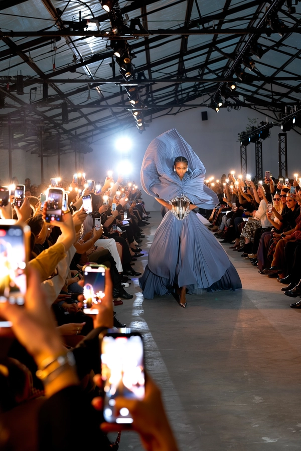 A commanding model halts abruptly mid-stride on the sleek runway, her voluminous dress caught in a dramatic swirl of stiff silk and tulle that seems frozen mid-motion, glittering under the cluster of flashing phones held aloft by enraptured guests. Her sharp gaze pierces through the halogen haze, framed by avant-garde sculptured shoulders and an intricate metallic corset that shimmers under the mix of cool blues and warm amber stage lights. The photogenic chaos of raised arms, blurred flashes, and whispering spectators lines the narrow catwalk inside a glass-paneled urban loft, the high ceiling crisscrossed with black truss beams and spot fixtures. The phone’s sensor captures subtle grain and a gentle bloom around the spotlight halos, enhancing the tactile textures of the runway’s scuffed concrete and the crisp pleats in her gown. Shot from an angled front-row perch with a slight horizon shift, the composition pulses with raw energy and edgy glamour—contemporary runway snapshot, shot on iPhone