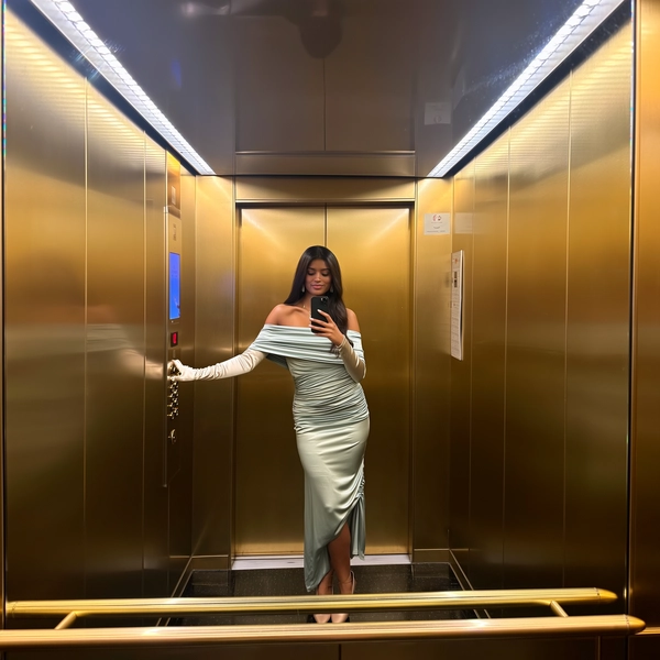 Rich gold panels envelope the elevator, their gleam multiplied by the mirrored ceiling and sides that fragment her image like a subtle prism effect. She stands serenely, draped in a fluid off-shoulder silk dress that shimmers gently under the soft cool LED strips overhead. The ruched fabric contrasts with the smooth satin sheen of her opera gloves, while nude ballet flats ground the look in effortless ease. Her gaze is fixed on the screen, face perfectly still, shadows and highlights tracing the realistic texture of her skin. The phone hovers at waist height, a faint fingerprint smudge settling on the glass as the doors inch closed behind her, her poised fingers barely touching the panel's brushed gold call button. The factory-fresh gloss of the elevator melds with subtle lens grain at the edges, framing an unbothered moment of quiet luxury—elevator mirror capture, urban chic on iPhone