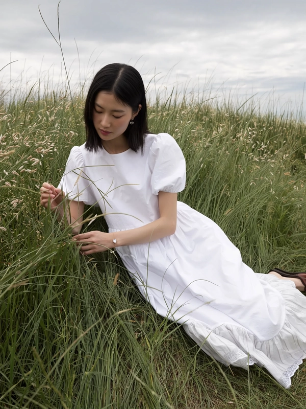 A gently contemplative moment captured as if on an iPhone, presenting a young woman reclining softly in tall grass beneath a cloudy sky. She wears a minimalist Jenny Fax-inspired white dress crafted from light cotton, subtly detailed with sheer puff sleeves and delicate scalloped lace trimming at the hem. Her straight dark hair is parted naturally, framing a serene, introspective expression enhanced by fresh, minimal makeup. The soft, diffused overcast light filters uniformly, accentuating the tender fabric textures and the gentle rustle of grasses. Surrounding her, the vintage-evocative meadow blends into a muted palette of soft greens and creams, while the natural layering of her dress offers an intimate tactile contrast. Framed slightly tilted and elevated, the composition balances elegant simplicity and wistful, casual charm, embodying Jenny Fax's whimsical femininity and quiet vintage romance.