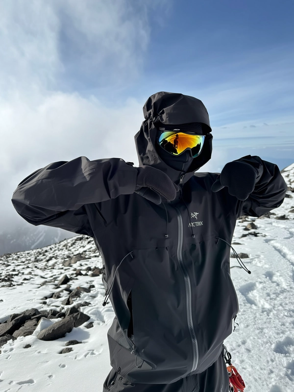 A candid, authentically spontaneous iPhone photo showcasing a person standing relaxed with arms gently lifted near the face amidst a jagged mountain ridge lightly covered with snow and patches of dark rock. The individual is dressed in an all-black Arc'teryx hooded waterproof jacket, hood pulled up and adjustable cords loosely hanging, exhibiting subtle fabric creases and realistic weathering from alpine conditions. Their head is covered by a matching black beanie partially visible under the hood, complemented by a functional, zippered face mask concealing the lower face. Reflective gold-tinted ski goggles cover the eyes, mirroring snowy peaks faintly visible in the blurred cloudy blue-gray sky background. Thick black gloves cover their hands, raised near the face in a natural gesture. Softly diffused natural daylight filtered by partial clouds casts sharp defined shadows on the snowy ground and rocks, accentuating the rugged textures of the technical gear and surrounding alpine environment. The composition is casually tilted and framed to reinforce the natural spontaneity and intimate feel of candid mountain photography, blending minimalist fashion with survival outerwear aesthetics synonymous with high-altitude winter explorations.