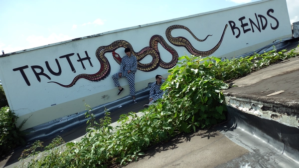 A candid iPhone-style street photo capturing two young men dressed in matching black-and-white checkerboard tracksuits, casually posing beneath a rooftop mural of cartoon-style snakes. The mural features prominently large, stylized graffiti text arranged as two separate lines reading:
"TRUTH"
"BENDS"
The snakes' warped and flowing shapes visually echo the vibe of the tracksuits. Surrounding the scene, lush green plants grow naturally out of weathered cracks in the concrete rooftop setting, adding authentic urban grit and life. Natural daylight casts subtle, contrasted shadows emphasizing the textures of peeling paint, worn concrete, and foliage. The framing is slightly off-center and tilted, maintaining an eye-level, spontaneous viewpoint typical of candid iPhone photography, capturing raw street authenticity combined with vivid graffiti culture aesthetics.