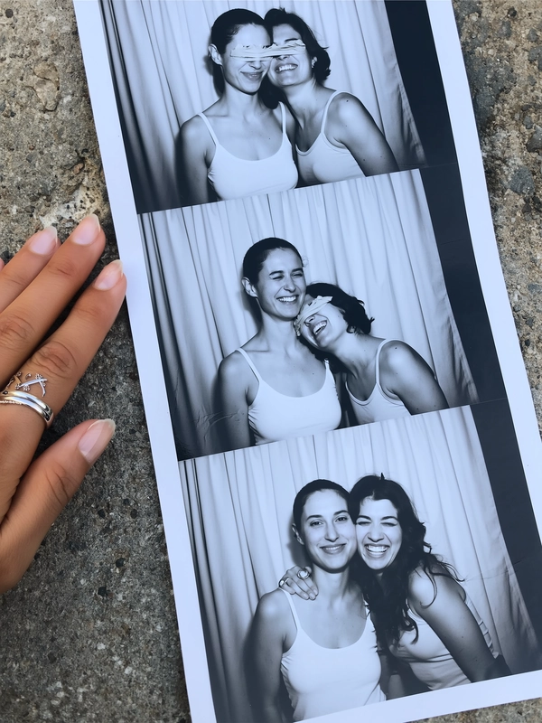 A vertical black-and-white photostrip lies casually on rough concrete near a hand adorned with silver rings, the contrast between the cool surface and monochrome tones enhancing authenticity. The strip showcases two women in simple tanks; in the first frame, one playfully covers the other's eyes, their smiles half-hidden and spontaneous. The following frame reveals both grinning fully into the lens, radiant and relaxed. Behind the subjects, a neutral curtain with faint folds and gentle texture creates the classic photobooth backdrop. The strip carries subtle creases and fingerprints, with soft shadows and muted highlights from a gentle iPhone flash. Slightly tilted and softly skewed, the composition feels like a genuine snapshot caught in an unguarded moment—contemporary photobooth snapshot, shot on iPhone.