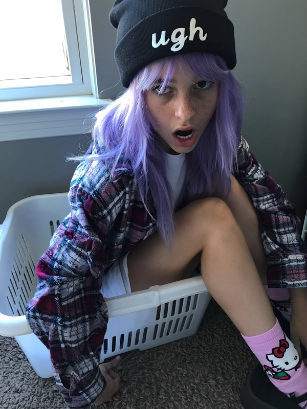 A candid casual snapshot of a teenage girl with vibrant lavender-dyed hair peeking from under a black beanie embroidered with "ugh." She sits inside a white plastic laundry basket in a softly lit bedroom, looking directly at the camera with an expression of exaggerated boredom. She wears a thrifted oversized flannel shirt layered over a white crop top, complemented by pastel pink Hello Kitty socks visible above chunky platform shoes. The ambient indoor light filters gently through a nearby window, producing muted daylight with intentionally softened contrasts and subdued saturation. Shadow regions show distinct cool-toned lavender-gray hues, emphasizing the mid-2010s photographic style. Textural details include visible individual hair strands loosely tousled beneath the beanie, realistic freckles and faint blemishes on her skin, detailed cotton fabric grain of the flannel with subtle wrinkles, and the fuzzy knit texture of the socks. The framing is casually informal with a slight tilt and off-center composition, reflecting spontaneous youthful snapshot authenticity typical of mid-2010s Tumblr-inspired candid photography.
