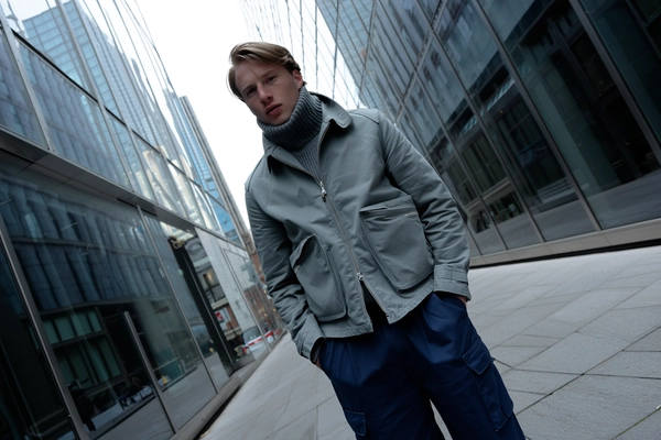 A candid iPhone-style capture of a pale-skinned man with light freckles standing in a cold urban alley flanked by reflective glass skyscrapers. He wears a slate-gray field jacket layered over a cozy turtleneck sweater and structured navy cargo pants, the textures of the fabrics and subtle skin details clearly visible. Soft, cool daylight reflects naturally off the glass facades, casting gentle ambient light and nuanced shadows. The framing is slightly tilted and asymmetrical, evoking a spontaneous, casual snapshot with natural posture and minimal self-awareness. The scene's overall tone is muted and cool, emphasizing genuine urban atmosphere and textural realism.