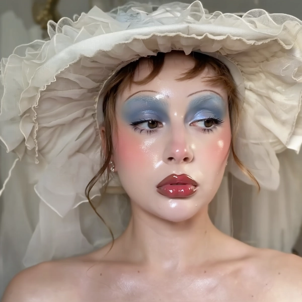 A close-up iPhone-style portrait of an elegant woman wearing a Victorian-inspired wide-brimmed hat with sculptural curves and delicate frayed edges, the textures of the fabric clearly visible and softly lit. Her skin has a dewy, glass-like finish with visible pores and a subtle natural glow across the nose and cheeks. Her makeup reflects a stylized babydoll aesthetic with an iridescent twist: deep blue eyeshadow softly blended across the lids with natural shimmer catching the light, heavy pink blush lightly dusted on her cheeks and below her eyes for a flushed effect, and glossy, vivid red lips reflecting warm studio light with a moist sheen. Her eyebrows are natural colored, softly brushed upward and unbleached, framing her expressive eyes. The photo is tightly framed from just above her head to below her collarbones, captured at a casual slight tilt to mimic a spontaneous handheld iPhone shot. The background is softly blurred with muted cream and pastel tones, hinting at vintage fabrics and subtle indoor textures without distraction. The lighting combines soft natural daylight with gentle phone flash highlights that enhance the gloss and shimmer on her makeup and dimensionality of her hat. Fine details like stray hair strands, fabric weave, skin texture, and lip gloss reflections are clearly visible, contributing to an intimate and authentic image that balances fantasy theatrical styling with grounded casual realism.