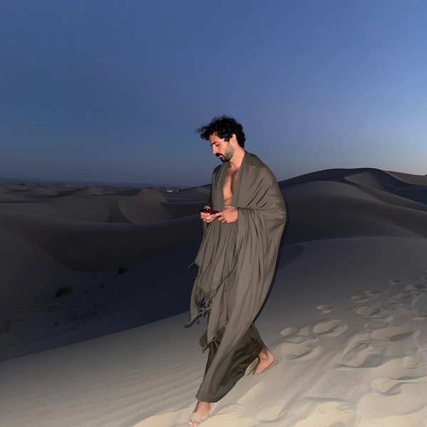 A candid, casually captured iPhone-style image of a Slovenian man dressed in muted desert tones, wearing a long, loosely wrapped shawl draped across his shoulders with subtle nomadic layering. He walks gracefully through expansive sand dunes at twilight, his silhouette elongated and subtly dramatic. The soft, low-contrast natural twilight light combined with the gentle glow of an iPhone flash creates a serene, introspective atmosphere with deep shadows and delicate highlights. The minimalist, slightly asymmetrical composition highlights the tactile textures of the flowing shawl fabric, the shifting sand, and the subtle skin nuances visible beneath the fabric. The scene evokes quiet elegance, mysterious allure, and the spontaneous authenticity typical of casual iPhone photography.