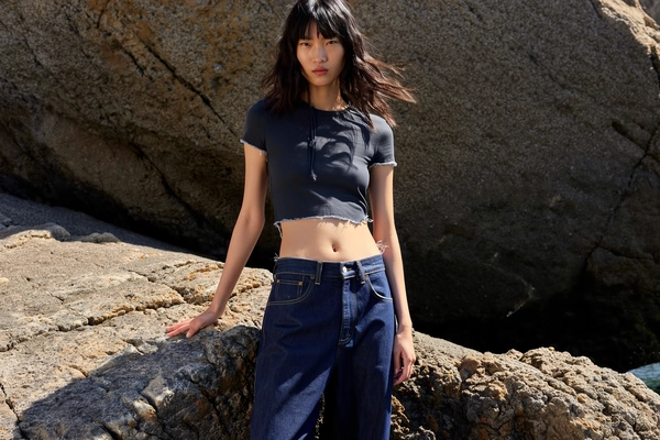 Midday sun fractures sharply across uneven jagged stones, casting deep chiaroscuro undercutting a cropped silk top and raw-edge denim tailored with deliberate fray—each fabric thread and rough indigo weave etched in vivid relief. The East Asian model stands statuesque, her gaze piercing forward with a quiet detachment that charges the stillness, lips parted ever so faintly, hair tousled naturally by a sea breeze that glosses skin with subtle chiaroscuro highlights.

Sunlight slices diagonally, a harsh key from above softened slightly by a golden fill to maintain detail in shadow-laced crevices of both stone and garment; deep shadows pool beneath the folds of distressed jeans, while the cropped top’s fine silk fibers catch and hold dappled gleams. Her expression twists the surrounding heat into inertia—an aloof statement echoing minimalist nonchalance.

Compositionally, the lens hovers at eye-level with a 50 mm focal balance, center framing the model as she occupies the ground’s fractured grid in crisp focus, edges yielding to a gentle vignette blur that frames without harshness. The jagged rocks stretch beyond, their stark textures seamlessly merging into shadow, amplifying the urban-wild contrast between structured denim and nature’s raw palette.

The image hums with a digital clarity reminiscent of sharp, film-like stillness where tactile grit and human presence fuse, retaining a subtle softness in the periphery to counterbalance the sharp sunlit crystal textures—high-fashion editorial, hyper-real texture fidelity