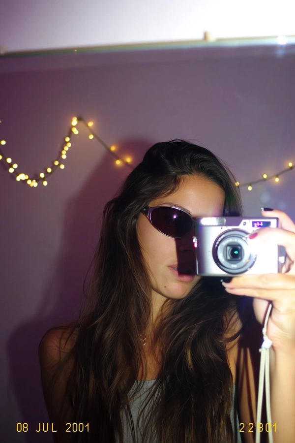 A quick pop from the built-in flash throws harsh shadows across her face, sunglasses reflecting crooked summer string lights glowing faintly behind her. Pixel fringing blurs the worn edges of the mirror she snaps into, and the silver digital camera peeks from her hand with a subtle flash bloom. CCD speckles dance in the pastel dimness, while the tungsten room light tugs warm highlights across the cool shadowed corners. The angle is slightly off-kilter, the soft focus edges stuttering as if the moment was caught mid-shake, timestamped “08 JUL 2001  22:45” in the bottom corner. The photo quality is low-resolution and somewhat grainy.—early-2000s digicam snapshot, grainy low-res quality
